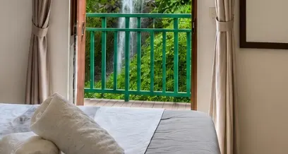 Puerta abierta a un balcón de baranda verde con la cascada al fondo; en primer plano, el borde de la cama con toallas.