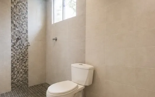 Baño privado con ducha de mosaico gris, sanitario blanco, paredes de cerámica clara y una ventana alta.