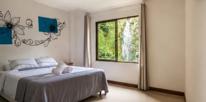 Habitación con cama doble, mural de flores con cuadros verde azulado y una ventana amplia por la que se ve la cascada entre la vegetación.