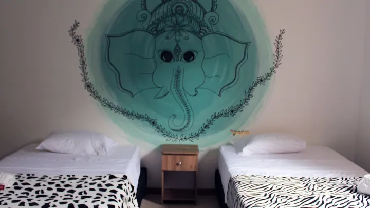 Dos camas con mantas de estampado animal bajo un mural de Ganesha verde agua, con una pequeña mesa de noche de madera.