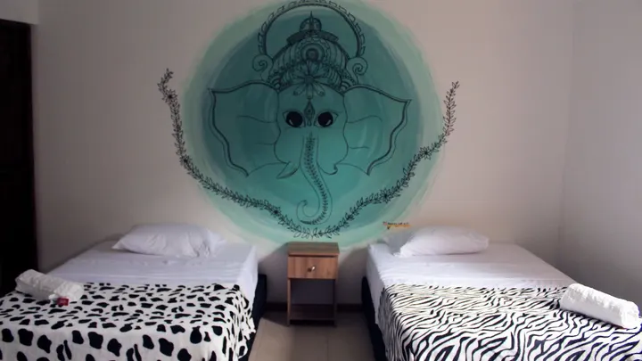 Dos camas con mantas de estampado de vaca y cebra bajo un gran mural de Ganesha pintado en verde agua, con una mesa de noche entre ambas.