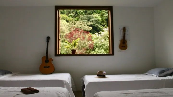 Habitación blanca con camas sencillas, una guitarra apoyada en la pared, un ukelele colgado y una ventana de madera abierta a árboles de follaje rojo y verde.