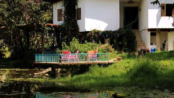 La casa blanca de dos pisos detrás del estanque, con una terraza de baranda azul y sillas rosadas sobre el agua.