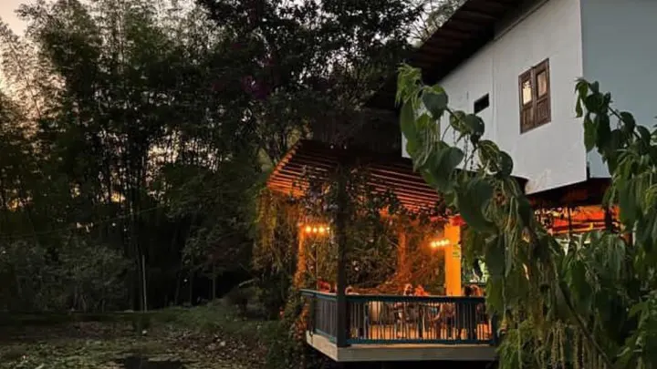 La casa al atardecer: luces cálidas encendidas bajo el alero de la terraza sobre el estanque, entre árboles oscuros.