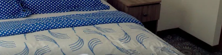 Cama con sábanas azules de puntos y edredón claro con estampado de ondas azules, mesa de noche de madera y alfombra de mosaico.