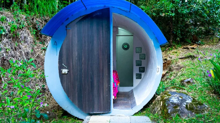 Cabaña tubo con techo azul y media puerta de madera abierta: dentro se ve el interior blanco curvo, una cama con manta fucsia y una puerta al fondo.