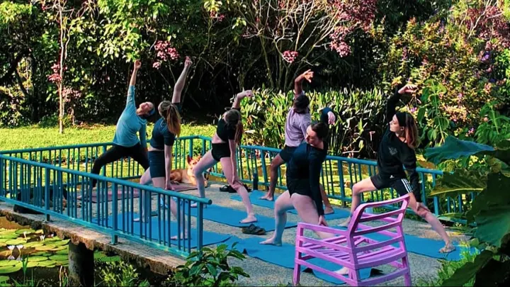 Grupo haciendo una postura de yoga con el brazo al cielo sobre la plataforma de baranda azul del estanque, junto a una silla fucsia.