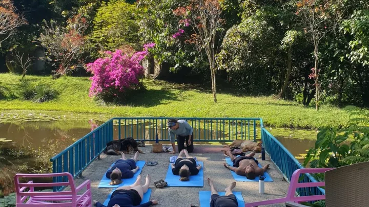 Personas en relajación final de yoga, acostadas sobre colchonetas azules en el deck del estanque, con buganvilias al fondo.