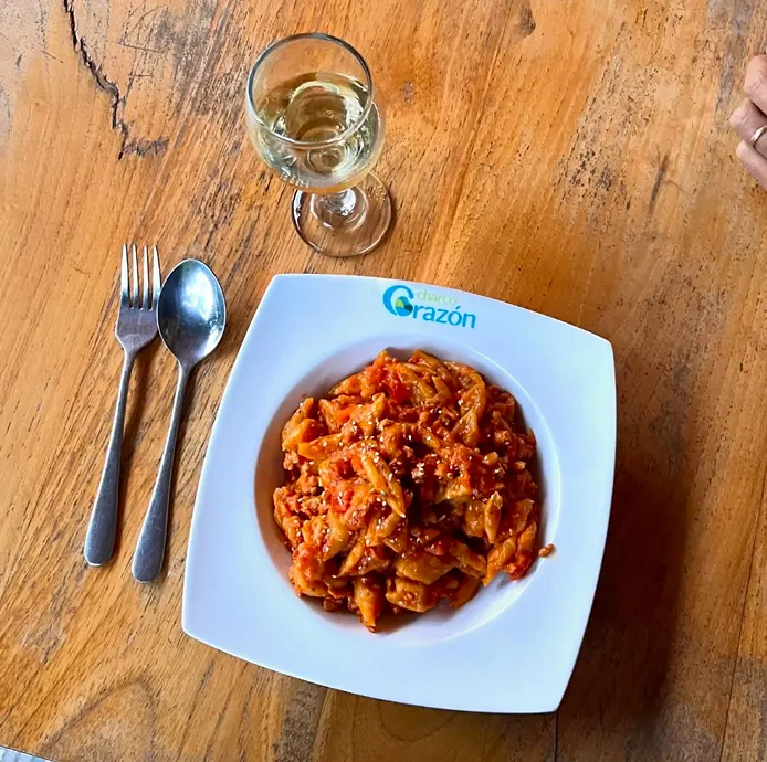 Plato cuadrado blanco con el logo de Charco Corazón, servido con pasta en salsa de tomate, junto a cubiertos y una copa de vino blanco sobre mesa de madera.