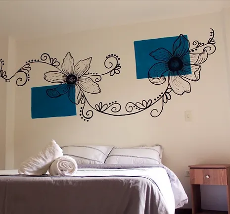 Cama doble con almohadas blancas bajo un mural de flores dibujadas en línea negra con cuadros verde azulado, junto a una mesa de noche de madera.