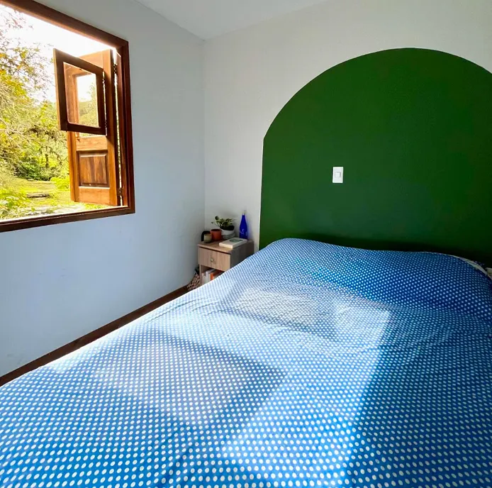 Habitación privada sencilla con un arco verde pintado como cabecero, cama doble con cubrecama azul de puntos y ventana de madera abierta al jardín.