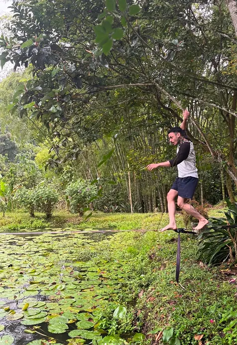 Un hombre descalzo hace equilibrio en una slackline a la orilla del estanque de nenúfares, bajo los árboles.