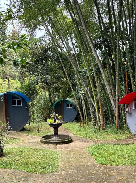 Tres cabañas tubo con techos azul, verde azulado y rojo, numeradas, entre guaduas altas y una pequeña fuente de piedra en el centro del sendero.