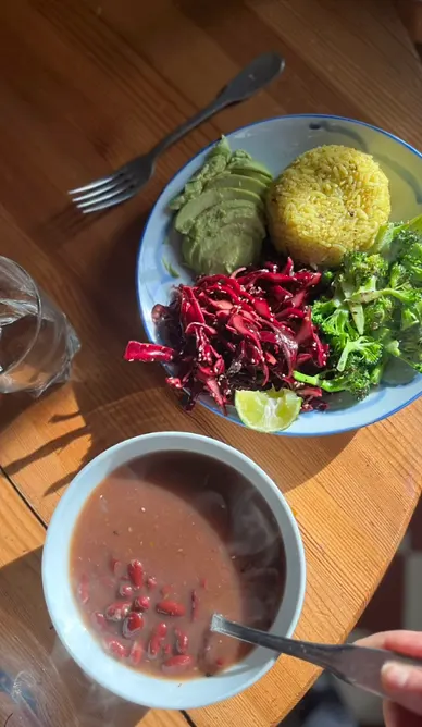 Plato vegetal con aguacate, torta de arroz amarillo, repollo morado, brócoli y limón, junto a una sopa de fríjoles rojos, sobre mesa de madera al sol.