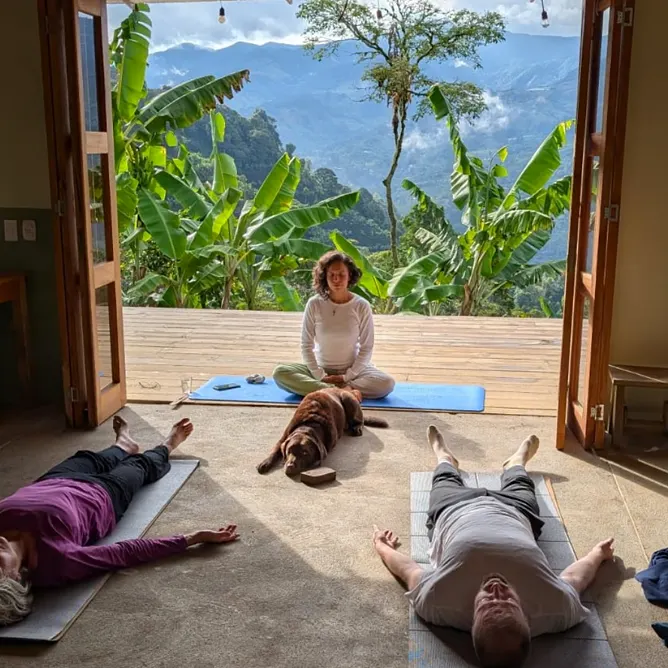 Clase de yoga junto a una puerta de madera abierta: una instructora medita frente a una terraza con plataneras y montañas, un perro marrón descansa y dos personas reposan sobre colchonetas.